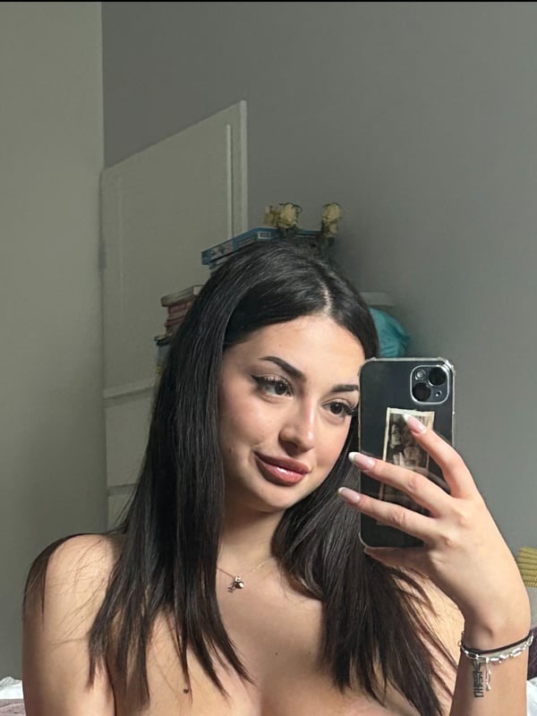 Young woman with long dark hair taking a close-up selfie in a bedroom, smiling softly with a phone in hand.