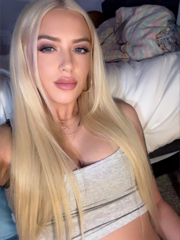 A close-up selfie of a young woman with long blonde hair and blue eyes, wearing a gray top, posing on a bed with a belly piercing visible.