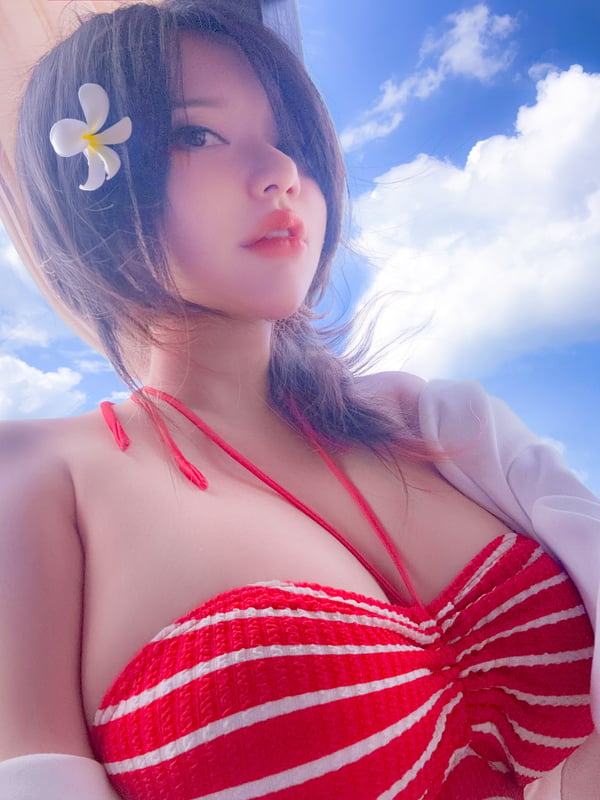 Young woman with dark hair and a flower in her hair, wearing a red striped swimsuit top, posing outdoors with a beach background.