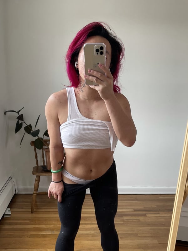 Person with pink hair taking a mirror selfie in a white tank top and black leggings, standing in a home interior with plants.