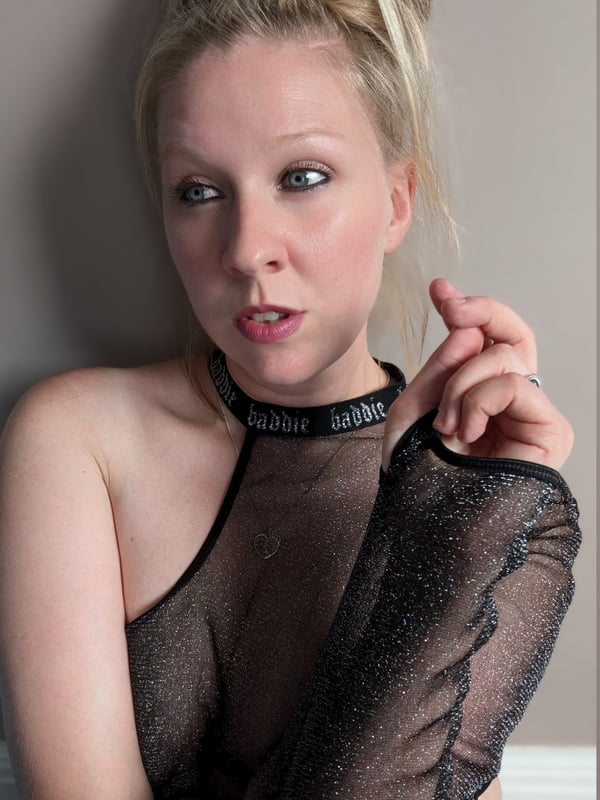 A woman with blonde hair in an updo, smiling with pursed lips against a wall, dressed in a sheer black mesh top.