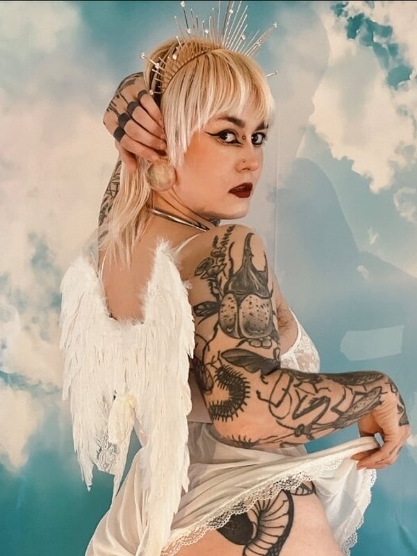 Person with blonde hair and tattoos poses sideways wearing white feathered angel wings and a flowing white outfit against a blue cloudy sky background.