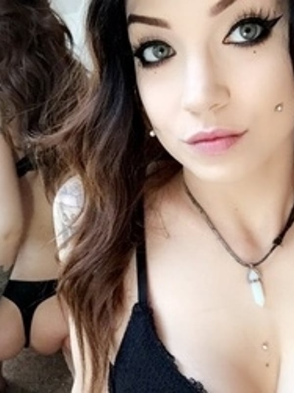 Close-up portrait of a woman with dark hair, piercings, and tattoos, wearing a choker necklace, with a mirrored reflection in the background.