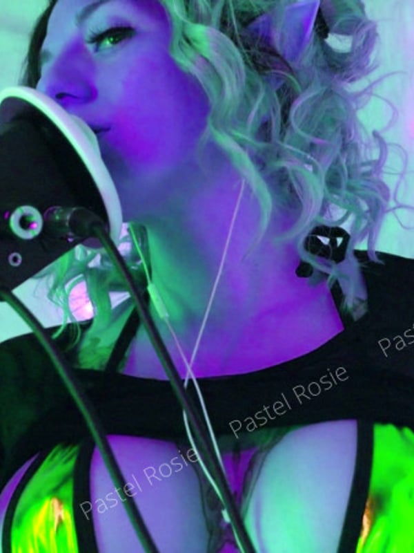 A model with dyed hair and tattoos posing playfully with a cup in a neon-lit room decorated with string lights.