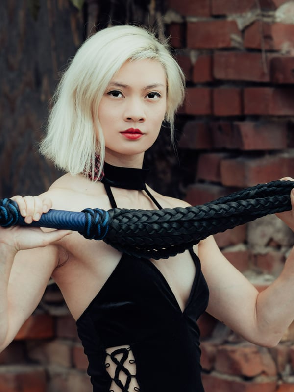 A woman with blonde hair holding a black flogger, dressed in a black lace dress against a brick wall background.