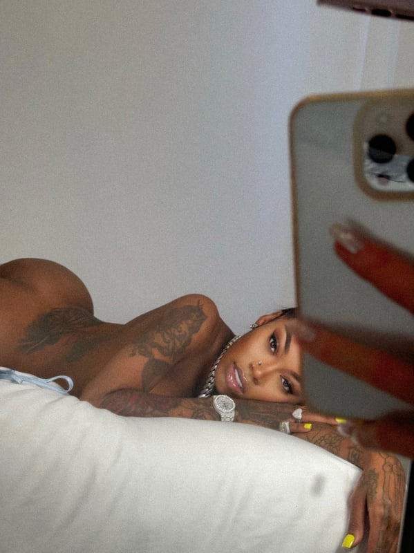 A woman with tattoos reclines on a bed holding a phone, posing confidently in a bedroom setting.