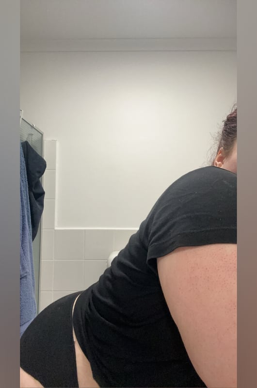A person with reddish hair posing sideways in a bathroom mirror, wearing a black top and shorts next to a shower.