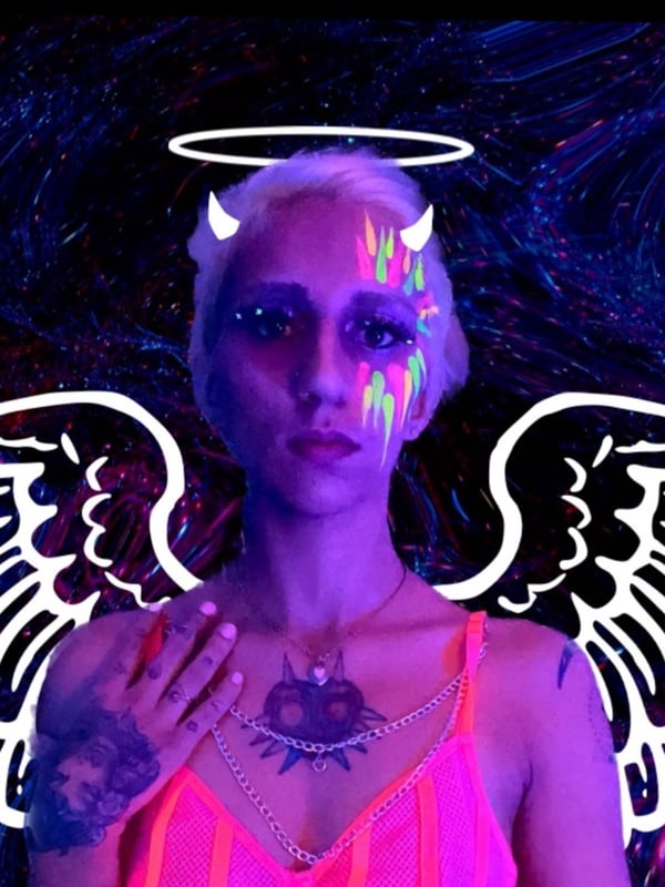 Woman with short blonde hair, devil horns, glowing face paint, and large white wings, posing in neon-lit setting wearing a pink top and jewelry.