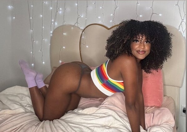 Young woman with curly hair posing on a bed in a colorful top and purple socks, with string lights and pillows in a cozy bedroom setting.