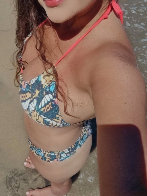 A smiling woman with wavy brown hair poses on the beach in a colorful bikini, standing with feet in the sand near the water.