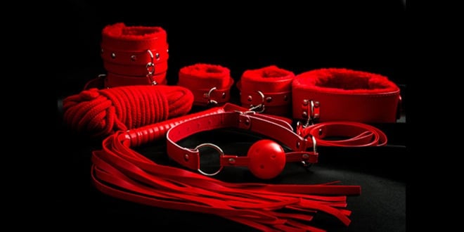 Arrangement of red leather bondage accessories including cuffs, ropes, collar, gag, and flogger displayed on a black background.