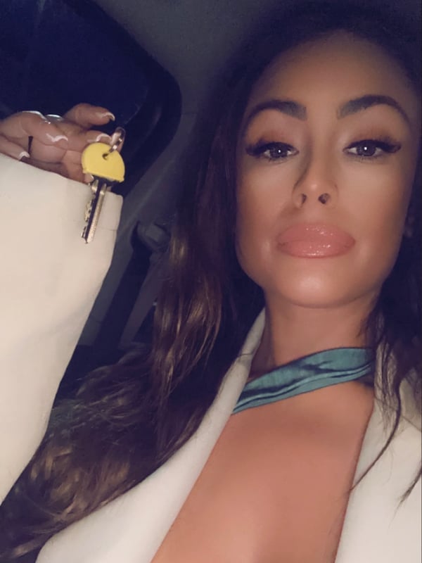 Close-up selfie of a woman with long wavy dark hair and bold makeup, wearing a white blazer, sitting in a car interior holding a yellow key.