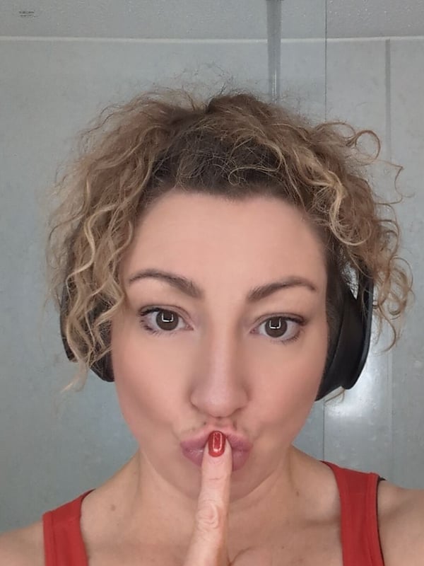 A woman with curly hair and headphones makes a playful shushing gesture with red lipstick, smiling in a casual indoor setting.