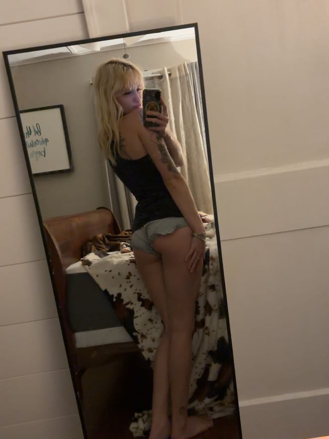 Young woman with long blonde hair taking a mirror selfie in a bikini top, holding a phone, with tattoos on her arm and a beach-themed background.