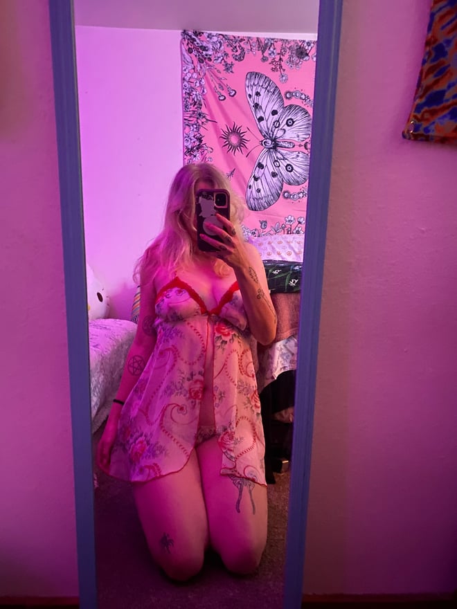 A woman with blonde hair kneeling in pink lingerie, taking a mirror selfie in a colorful bedroom with neon lighting and patterned wall hangings.