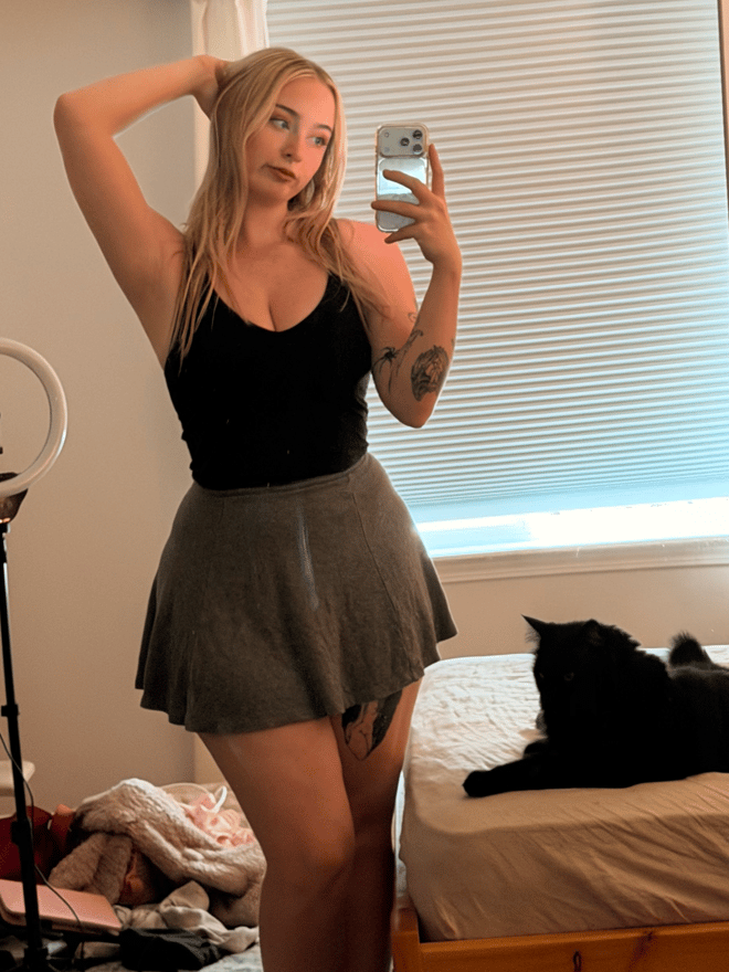 A blonde woman with winged eyeliner poses in a light-colored top, with visible tattoos and a nose piercing.