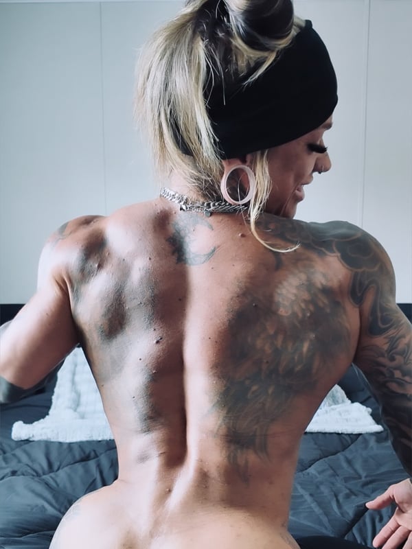Muscular woman with extensive tattoos on her back, blonde hair in a ponytail, wearing a black headband and choker with piercings, posing confidently indoors.