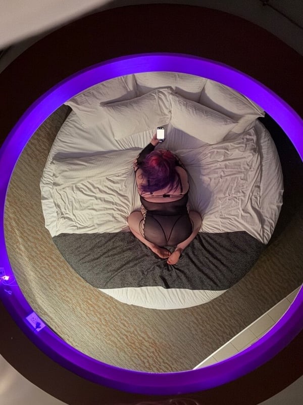 Overhead view of a person with colorful hair sitting on a bed in a dimly lit room with purple lighting and white sheets.