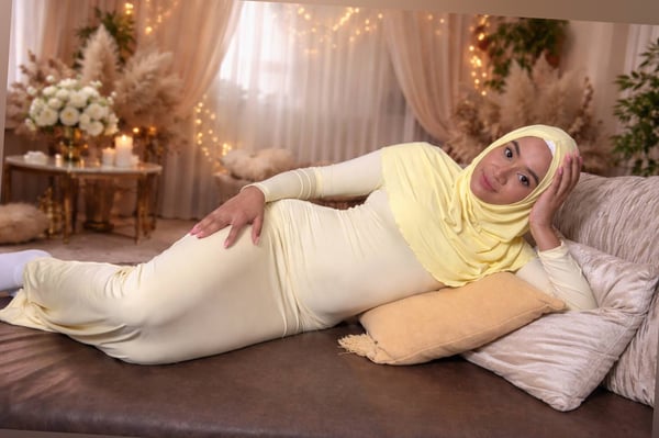 Woman in a yellow hijab and long dress reclining on a couch in a decorated room with plants, candles, and sheer curtains.