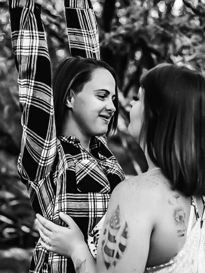 Two women embracing and smiling in a black and white photo, wearing plaid shirts with arms raised in a forested setting.