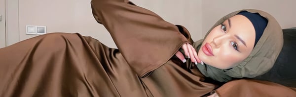 A woman wearing a hijab and flowing brown outfit poses reclined on a bed with a thoughtful expression.