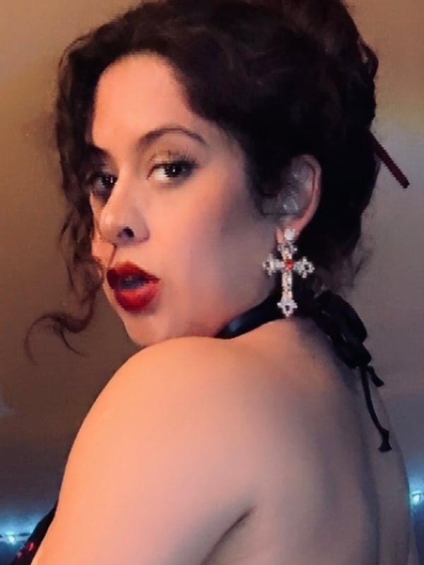 Close-up portrait of a woman with curly dark hair, red lips, and large earrings, wearing a black choker against a warm background.