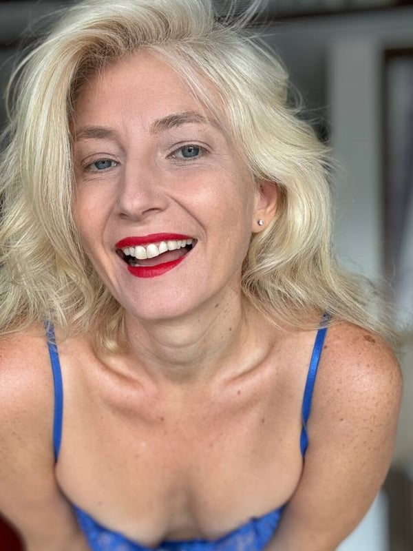 A smiling blonde woman with long hair wearing a blue top, posing indoors.
