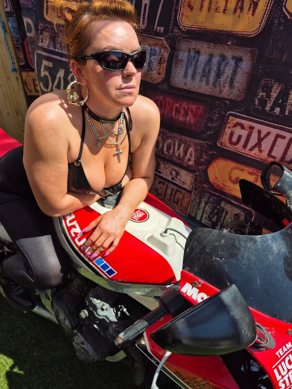A woman with red hair wearing sunglasses and a choker necklace, posing on a red motorcycle against a graffiti wall background.