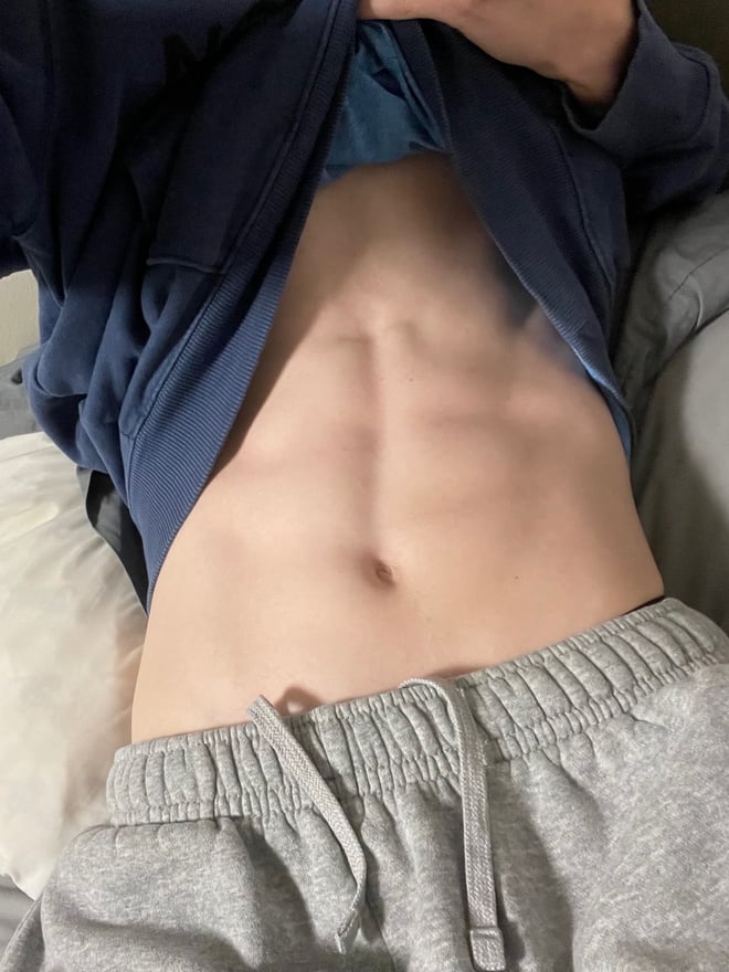 A young man taking a selfie on a bed, hoodie open showing defined abs, wearing gray sweatpants.