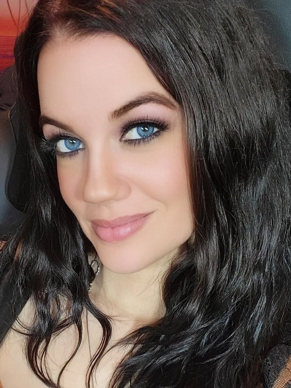Woman with long wavy dark hair and blue eyes smiling in a gaming chair at sunset, wearing a lace top.