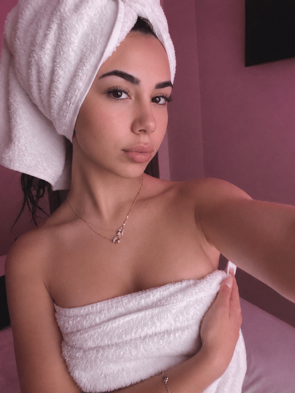 Young woman with long dark hair taking a mirror selfie in a bedroom, wearing a light pink tank top and white bottoms while kneeling on a fluffy pink rug.