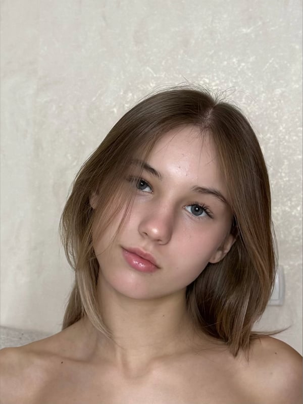 Close-up portrait of a young woman with long brown hair, gazing sideways with a subtle smile in a softly lit room.