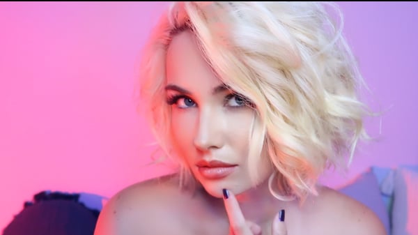 A young woman with blonde wavy hair posing confidently with a subtle gesture against a vibrant pink and purple background.