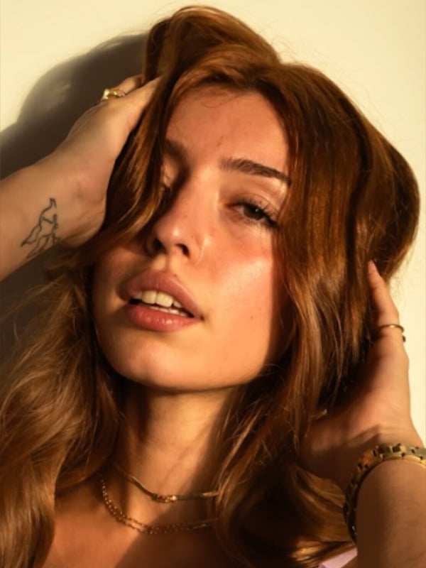 A close-up portrait of a person with long brown hair posing with hands in their hair.