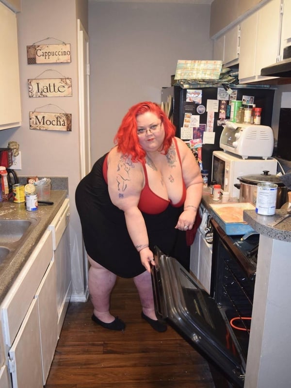 A woman with red hair stands in a kitchen, wearing a black top, opening an oven while surrounded by kitchen appliances and utensils.