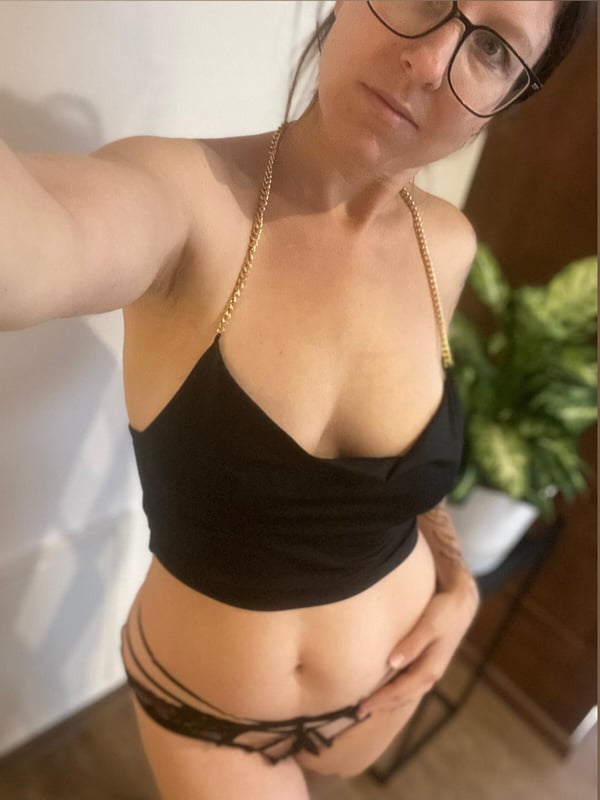 A woman taking a selfie in a black top and bottoms, wearing glasses, with a plant in the background.