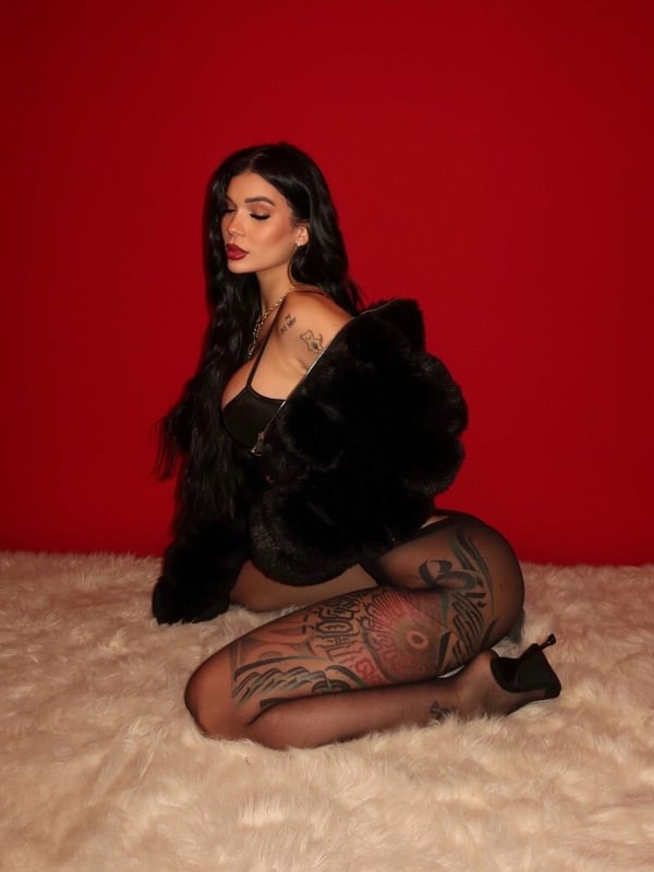 A woman with long dark hair sits on a fur rug against a red background, dressed in black attire and heels, showcasing tattoos.