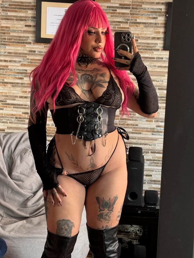 A blonde woman with dramatic makeup, facial piercings, and extensive tattoos posing indoors in dark alternative-style clothing.