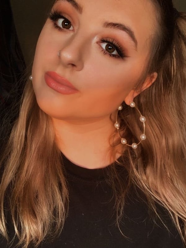 Close-up portrait of a young woman with blonde hair in pigtails, wearing hoop earrings and a black top, smiling softly against an indoor background.