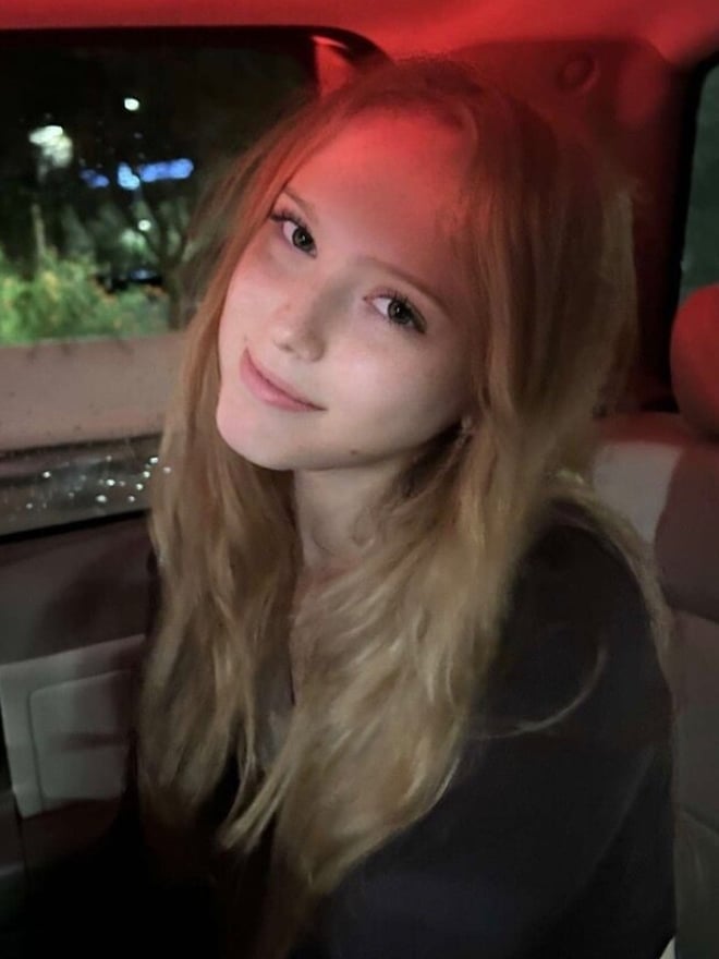 A young woman with light hair smiling from inside a car at night.