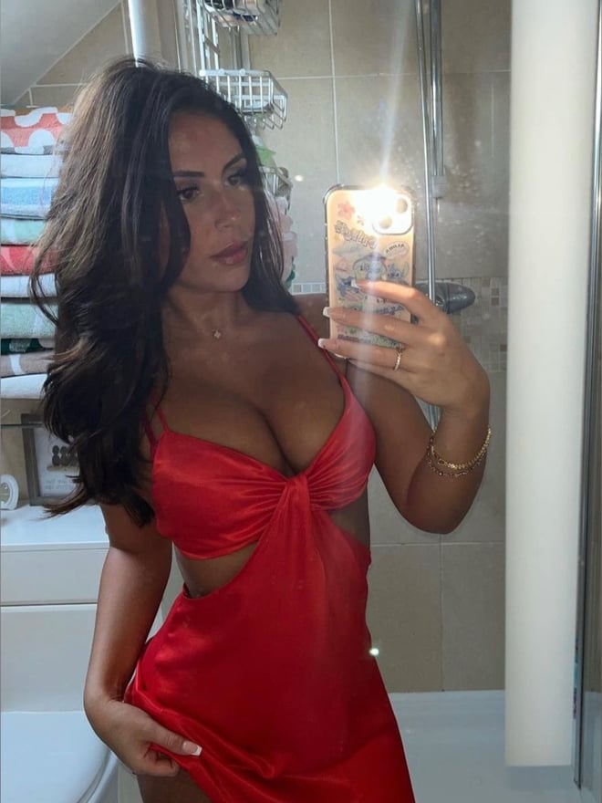 A woman in a red cut-out dress taking a mirror selfie in a bathroom with a shower.