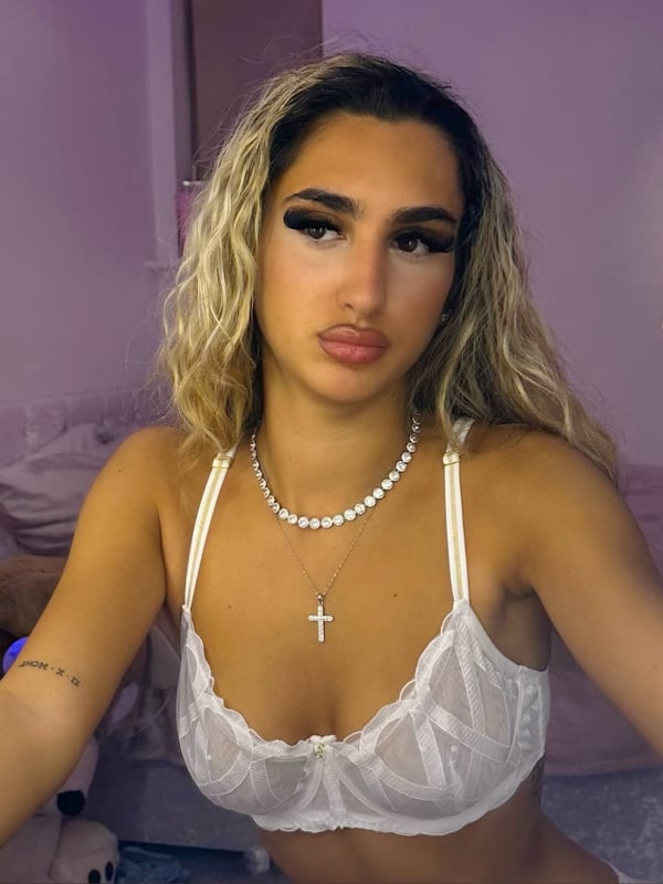 A young woman with wavy blonde hair taking a close-up selfie in a lace top, holding her phone in a purple-lit bedroom.