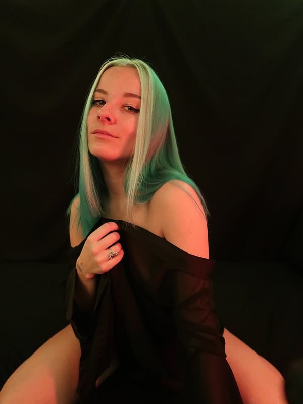 A woman with teal hair smiling at the camera, wearing an off-shoulder black top against a dark background.