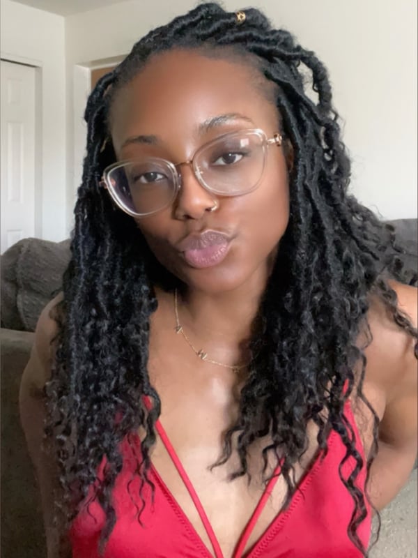 Young woman with long braided hair, wearing glasses and a red top, posing playfully indoors.