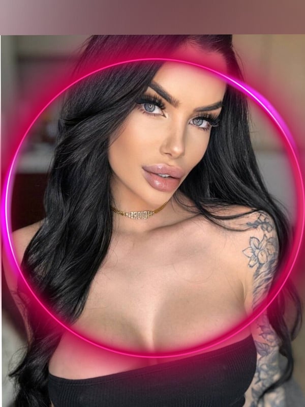 Close-up portrait of a woman with long dark hair, striking makeup, and tattoos on her shoulder, framed in a glowing pink circle against a dark background.
