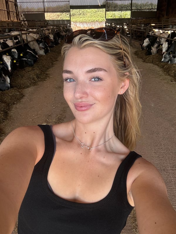 A smiling blonde woman taking a selfie in a barn with cows and wearing sunglasses and a black top.