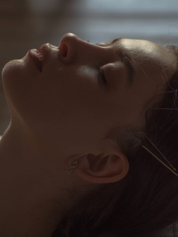 A young woman with long dark hair reclining with her head tilted back and eyes closed, wearing small earrings, in a softly lit indoor setting.