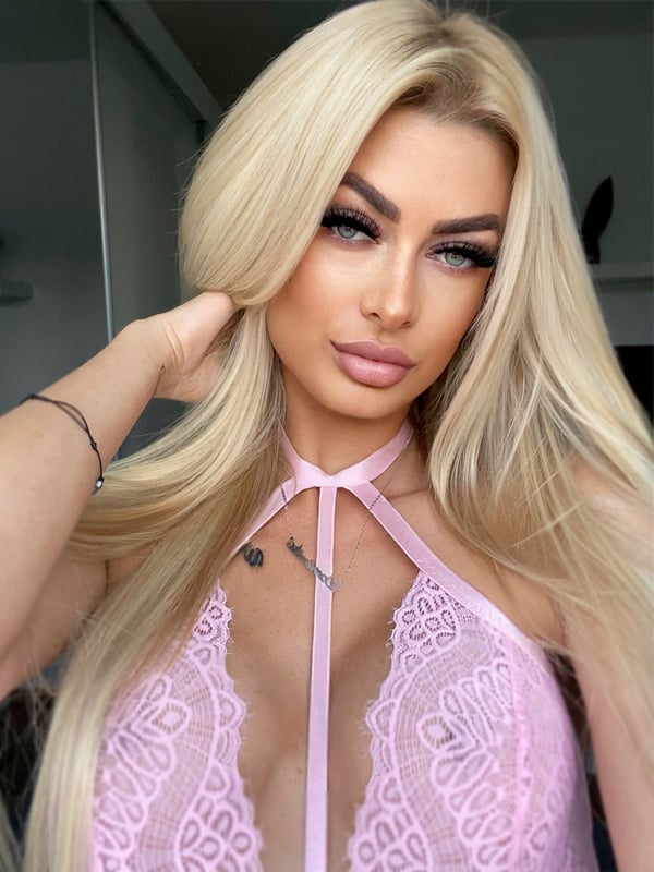 A close-up of a woman with long blonde hair and bold makeup, posing in a pink lace top.