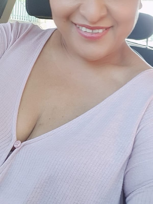 Close-up portrait of a smiling woman with long dark hair, wearing a pink top, seated in a car.