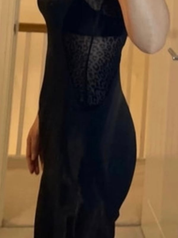A woman standing indoors, posing confidently in a form-fitting black sleeveless dress with lace details.